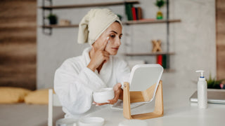 Woman in robe and towel applying skincare cream in front of mirror