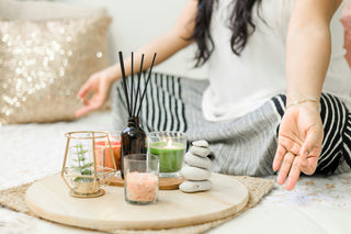 A meditation setup featuring a round wooden tray with lit candles, a reed diffuser, pink salt crystals, a small plant in a golden holder, and stacked smooth stones. In the background, a person sits cross-legged, creating a peaceful and calming atmosphere.